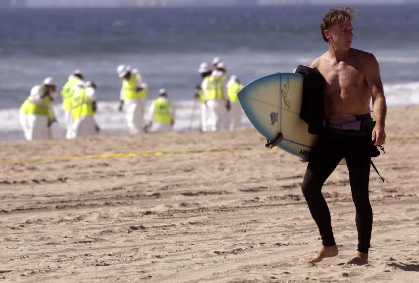 HUNTINGTON BEACH, CA - OCTOBER 5, 2021 - - Surfer Eddie McKee walks near workers who continue cleaning the beach from a recent oil spill near the Huntington Beach Pier on Saturday, October 5, 2021. He was sizing up the situation to see if it was safe to surf. McKee runs Salt Water Tacky surf wax business from Huntington Beach. (Genaro Molina / Los Angeles Times)