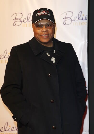 A man posing in a black coat, tinted glasses and an "Obama" baseball cap