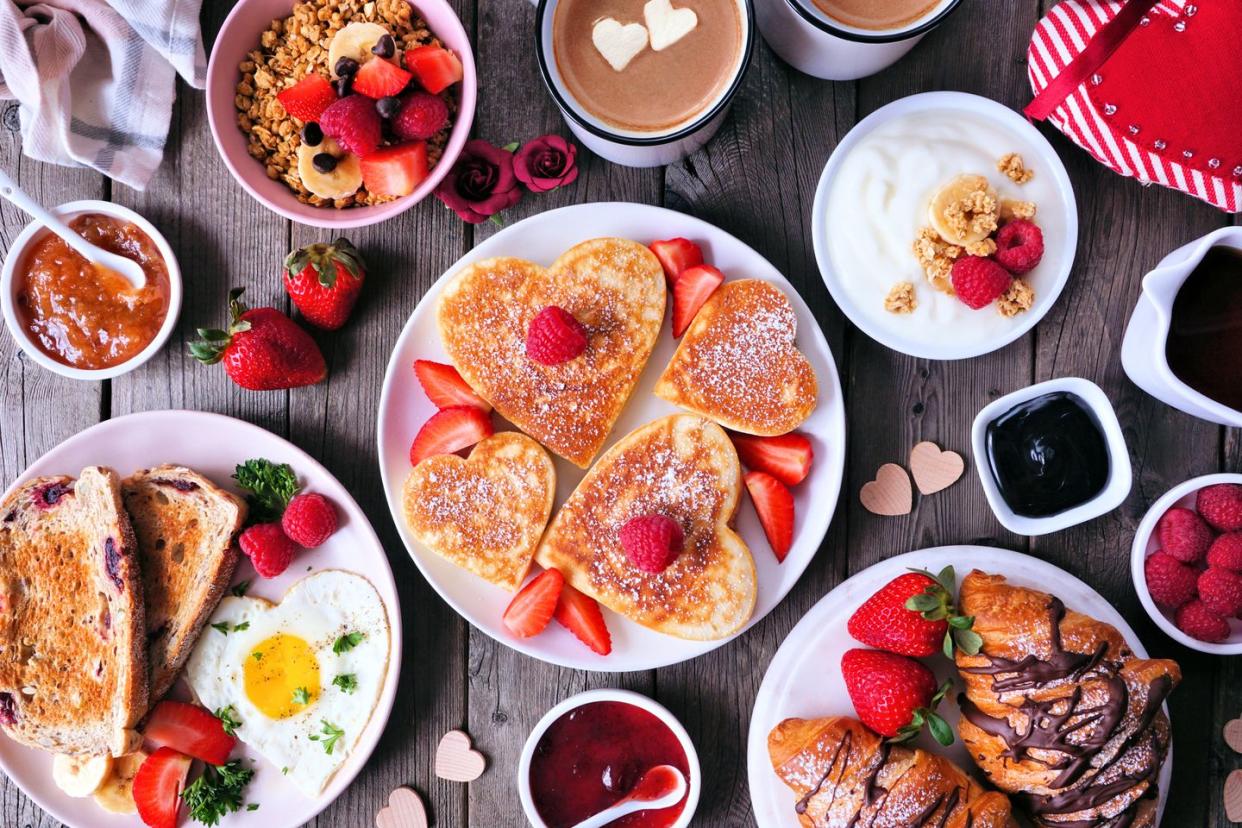 valentines or mothers day breakfast table scene on a dark wood background with heart shaped pancakes, eggs and love themed food