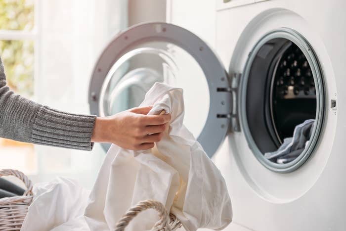 A person loading a white sheet from a basket into the washing machine. The image is probably related to household chores or laundry tips.