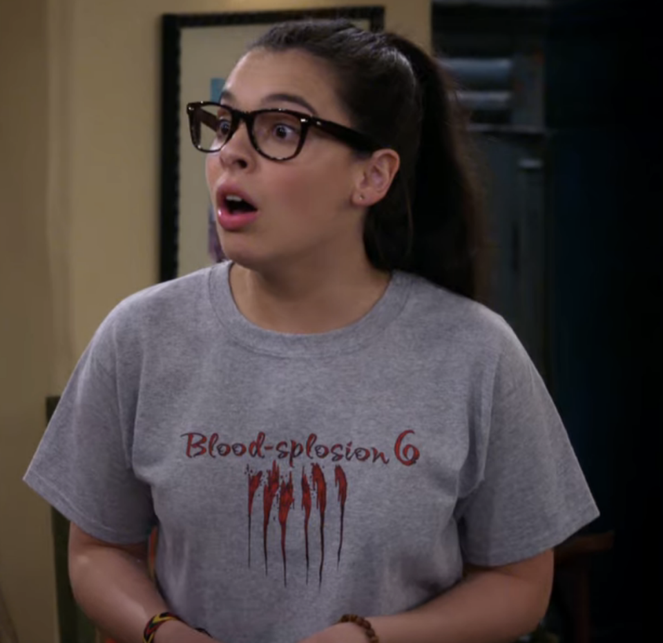 Isabella Gomez, with long hair tied back, wearing glasses and a "Blood-splosion 6" T-shirt, looks surprised with her mouth open