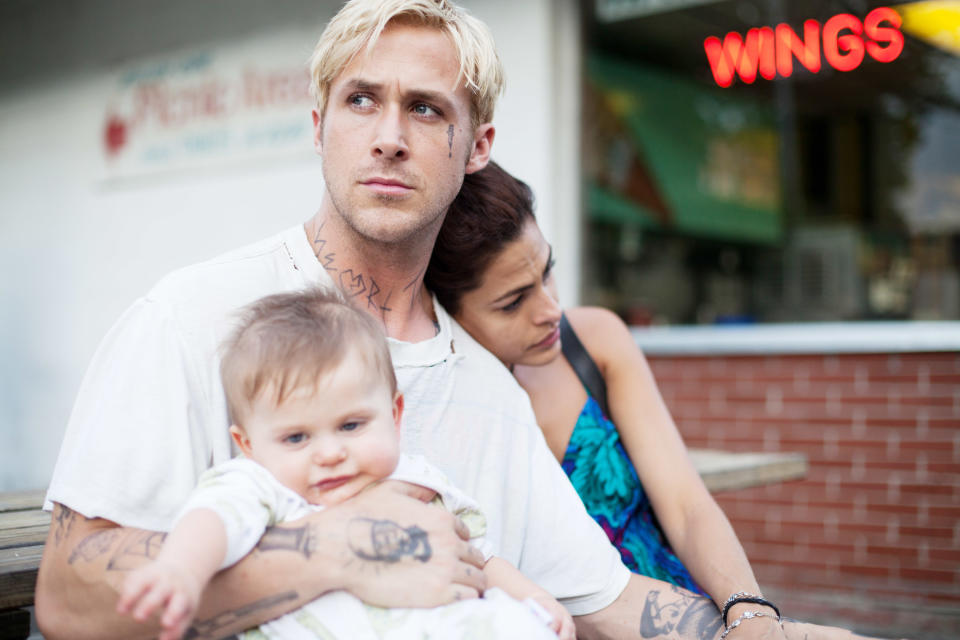 Ryan Gosling, seated and tattooed, holds a baby while Eva Mendes leans on him in front of a diner with a "WINGS" neon sign