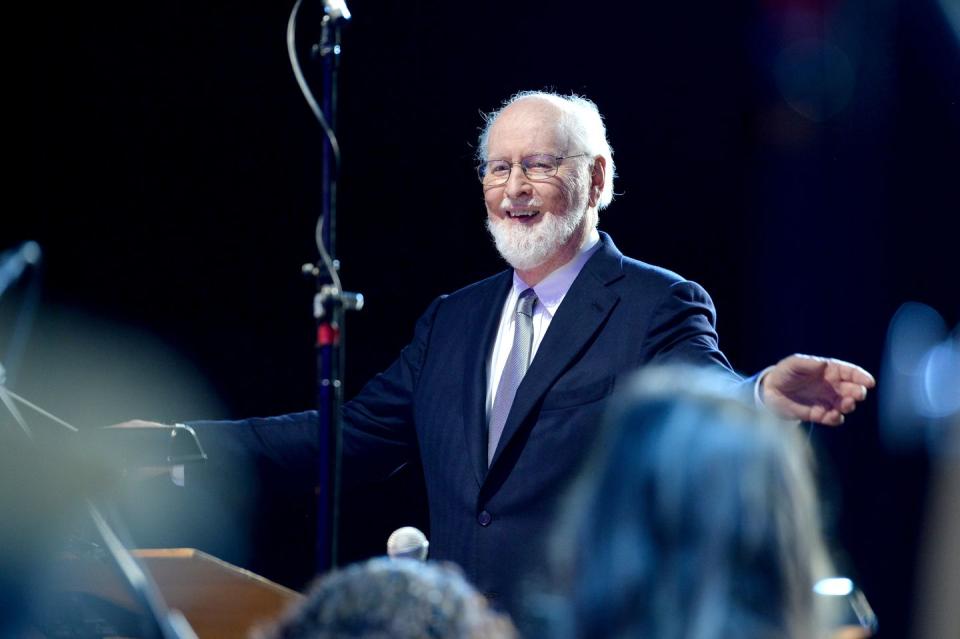 composer john williams holding his arms out to lead an orchestra and smiling