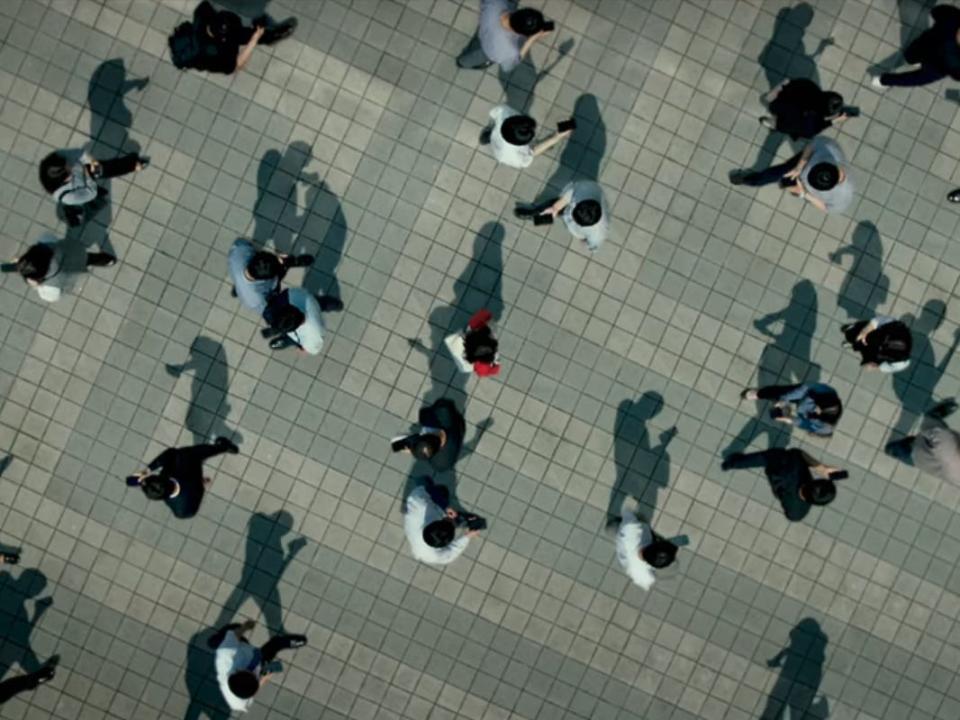 na-mi in unlocked stands still in the center of a busy pedestrian area. she's wearing red, while everyone around her is wearing neutral colors and walking around while using their phones