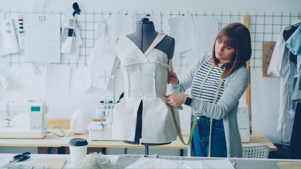 young seamstress is measuring clothing pinned to tailoring dummy with measure tape attractive woman is concentrated and thoughtful studio is light, modern, with many sewing tools and items