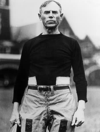 An older John Heisman is dressed in a football uniform, including a dark knit top and padded pants.