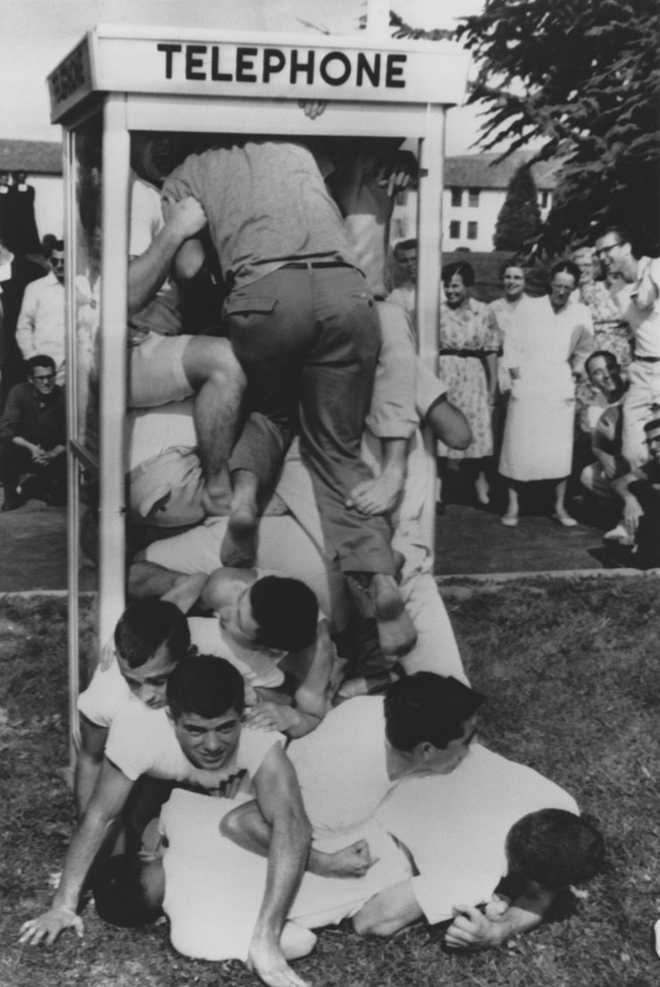 A large group of men, many dressed similarly, are attempting to fit inside a telephone booth while onlookers watch and laugh in the background