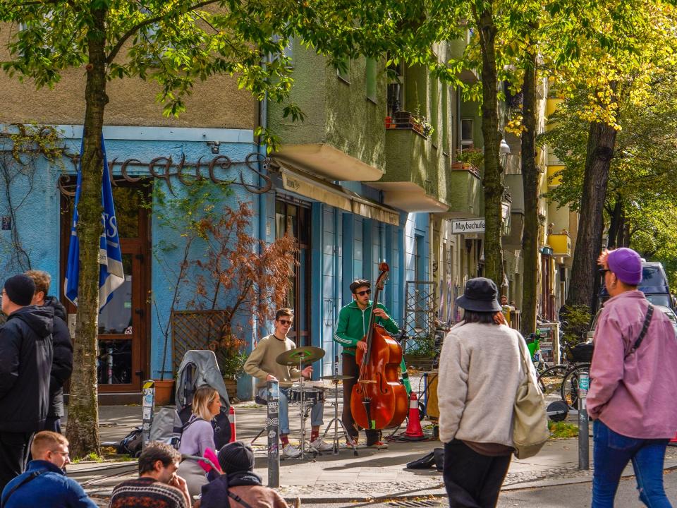 Musicians play on a lush street corner with pedestrians sitting and walking by.