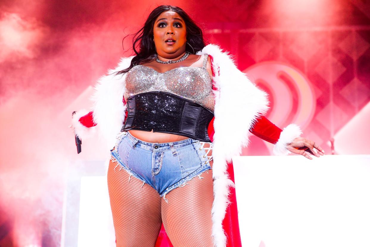 Lizzo stands on stage wearing a rhinetsone corset, jean shorts and a Santa Claus robe