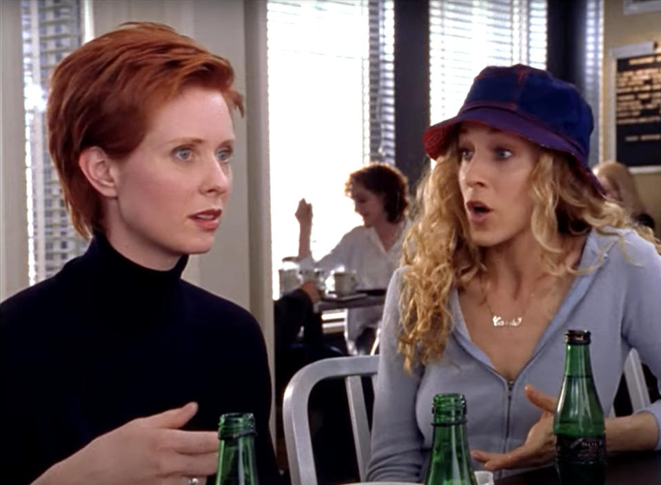 Cynthia Nixon and Sarah Jessica Parker are sitting at a café table in conversation. Both have bottles of sparkling water in front of them