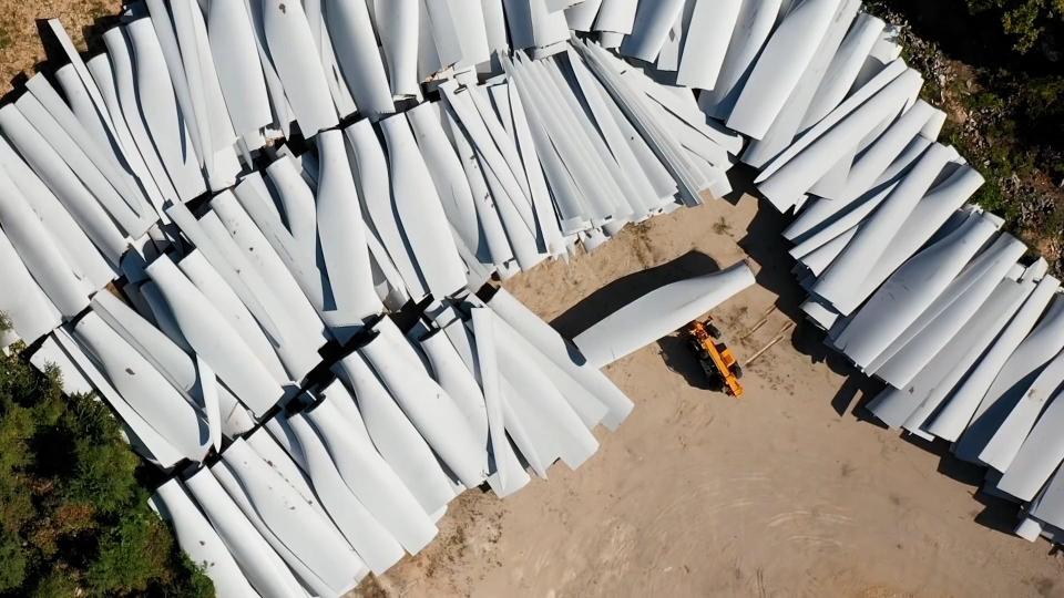 Aerial view of used wind turbine blades stacked on the ground. The rows of blades extend out of frame on each side.