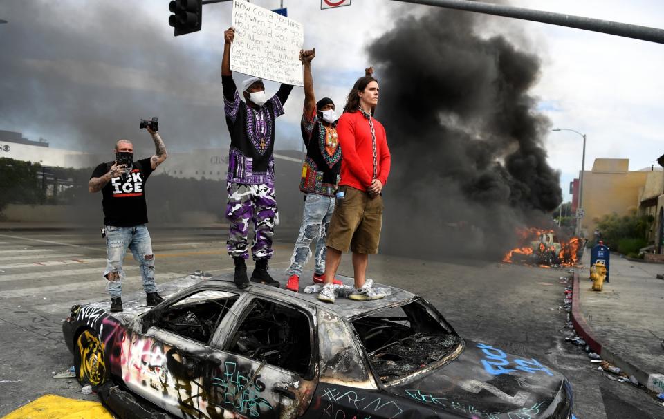 Protesters stand atop a burned, graffiti-covered LAPD cruiser. Flames rise from another cruiser nearby.