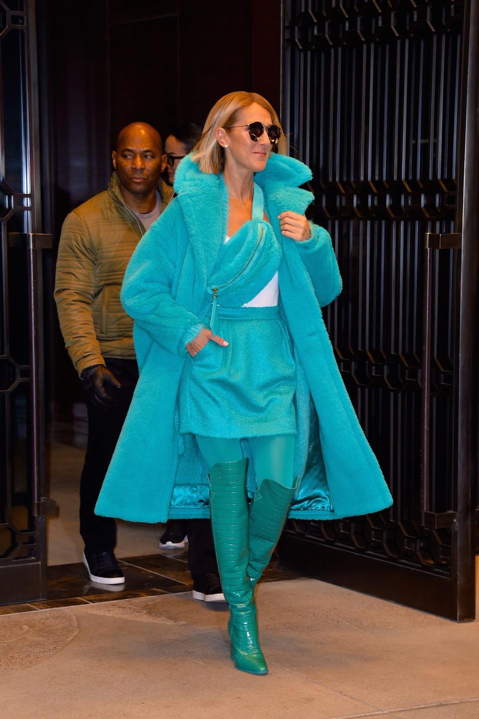 Celine in a turquoise blue furry coat with a matching fanny pack worn across her chest. She's also wearing a turquoise skirt, tights, and over the knee boots.