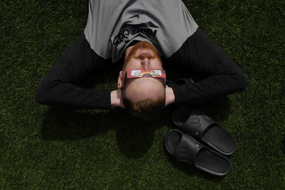 Chicago White Sox pitcher Steven Wilson lays in the grass outside the dugout and looks up through special glasses during a total solar eclipse, at Progressive Field in Cleveland on Monday, April 8, 2024, before the Cleveland Guardians home opener baseball game against the White Sox. (AP Photo/Carolyn Kaster)