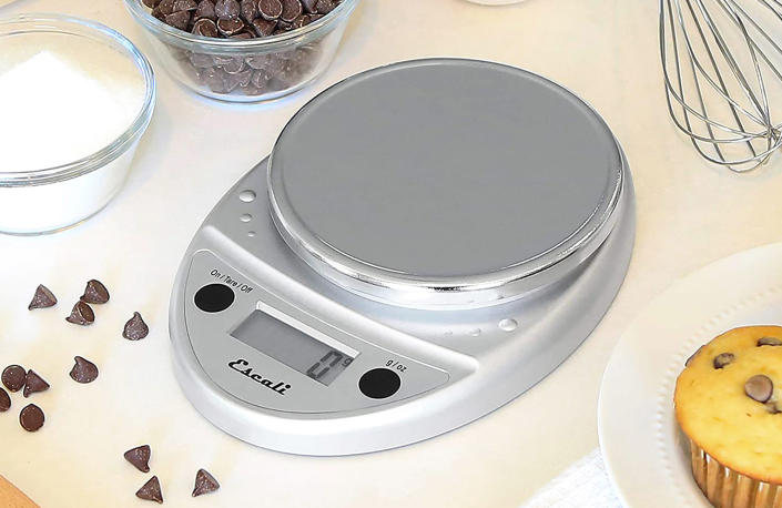 The Escali Primo food scale on a table surrounded by baking supplies including a bowl of sugar, a bowl of chocolate chips and a muffin.