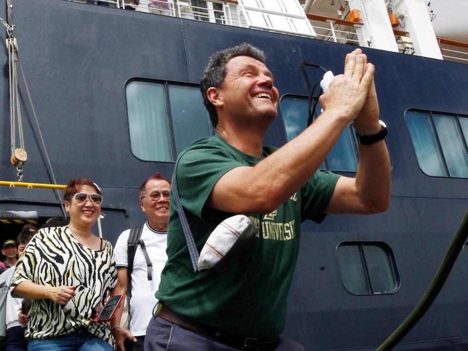 ADDS NAME OF PASSENGER - FILE - In this Feb. 14, 2020, file photo, John Miller, from Orcas Island, Wash., reacts after he disembarked from the MS Westerdam, back, at the port of Sihanoukville, Cambodia. Having finally reached a friendly port willing to accept them and stepped ashore after weeks of uncertainty at sea, hundreds of cruise ship passengers eyed warily over fears of a new virus are now simply trying to find a way home. (AP Photo/Heng Sinith, File)