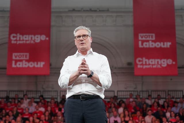 Sir Keir Starmer with hands clasped, speaking on a stage with Labour supporters and banners behind him