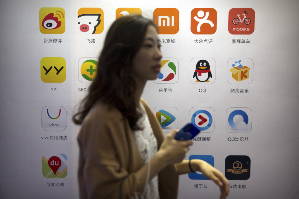 A woman holds her cellphone as she walks past a display showing the icons of popular Chinese smartphone apps at the Global Mobile Internet Conference (GMIC) in Beijing, Thursday, April 26, 2018. The GMIC features current and future trends in the mobile Internet industry by some major foreign and Chinese internet companies. (AP Photo/Mark Schiefelbein)