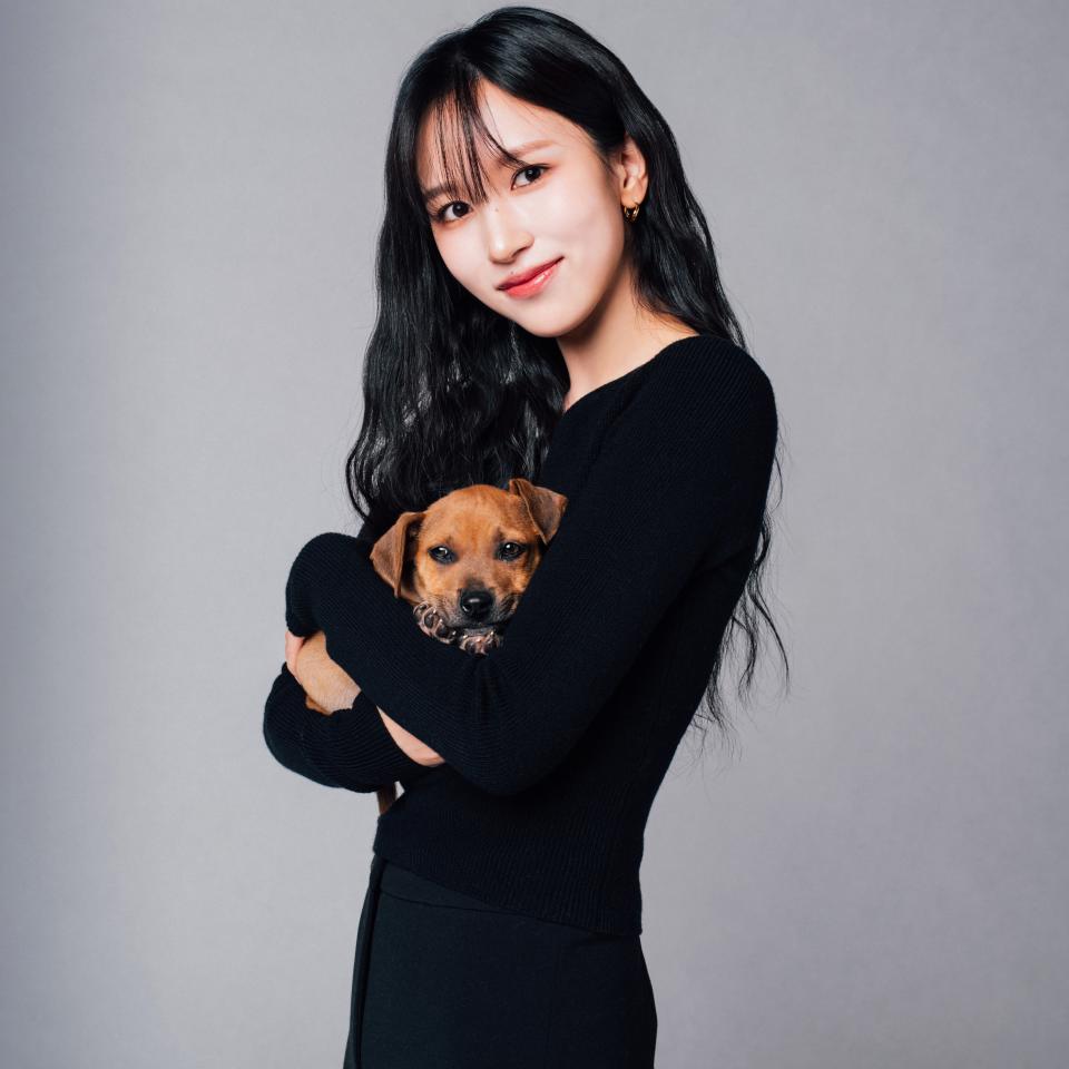 Woman in a black outfit smiling, holding a small puppy against a neutral background