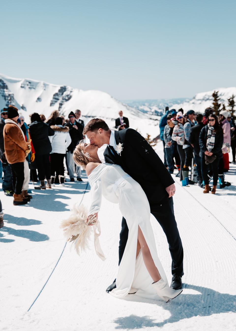 A groom dips and kisses a bride on a snowy mountain.