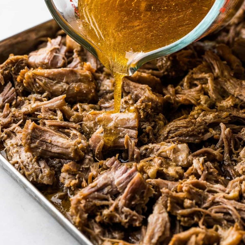 A glass measuring cup pouring liquid over shredded pork in a baking tray