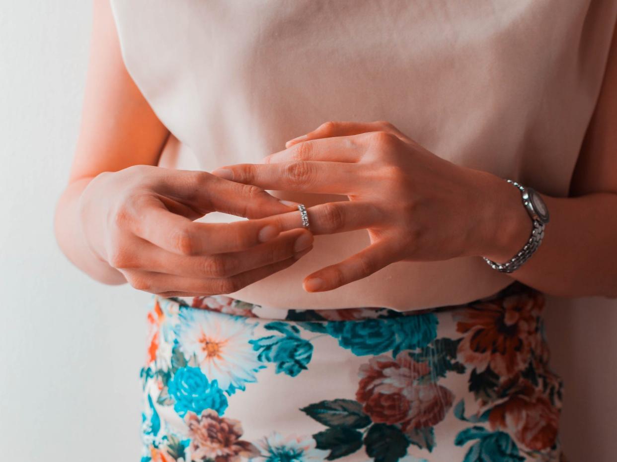 Image from the chest down of a person presenting as a woman taking off her diamond wedding ring. She wears a silver watch on her left hand, a white sleeveless top, and a white skirt with a blue, white, and pink pastel floral pattern.