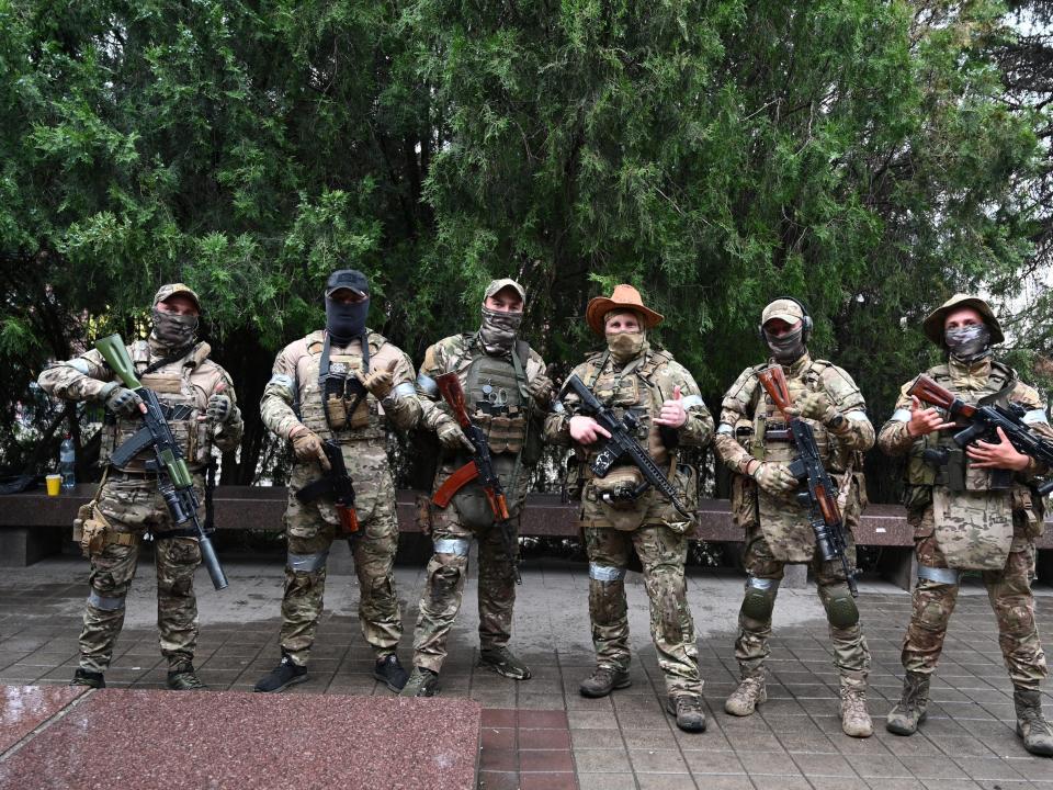 A row of six soldiers in uniform and ski masks holding guns on a brick road