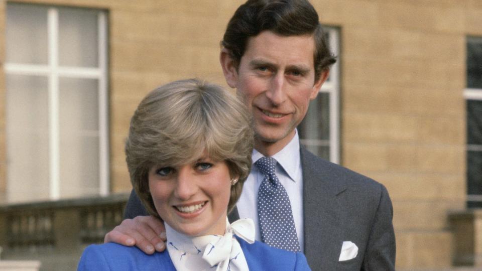 prince charles and lady diana spencer stand on concrete steps and smile for photos, she holds a black purse, he rests a hand on her shoulder