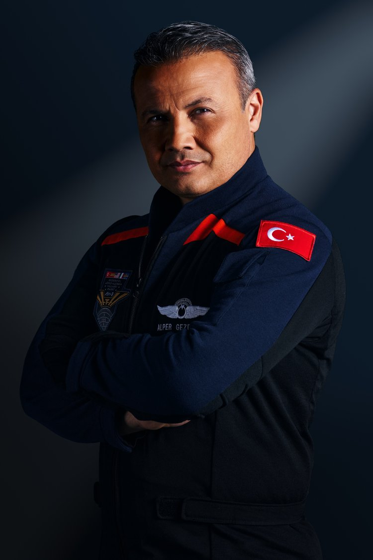 A man stands with crossed arms, wearing dark blue jumpsuits with red accented shoulder wings.  He looks serious.