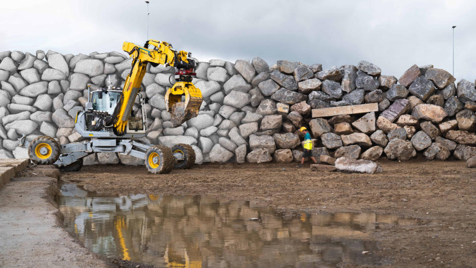     A yellow excavator holds a large boulder, part of its project to build a rock wall. 