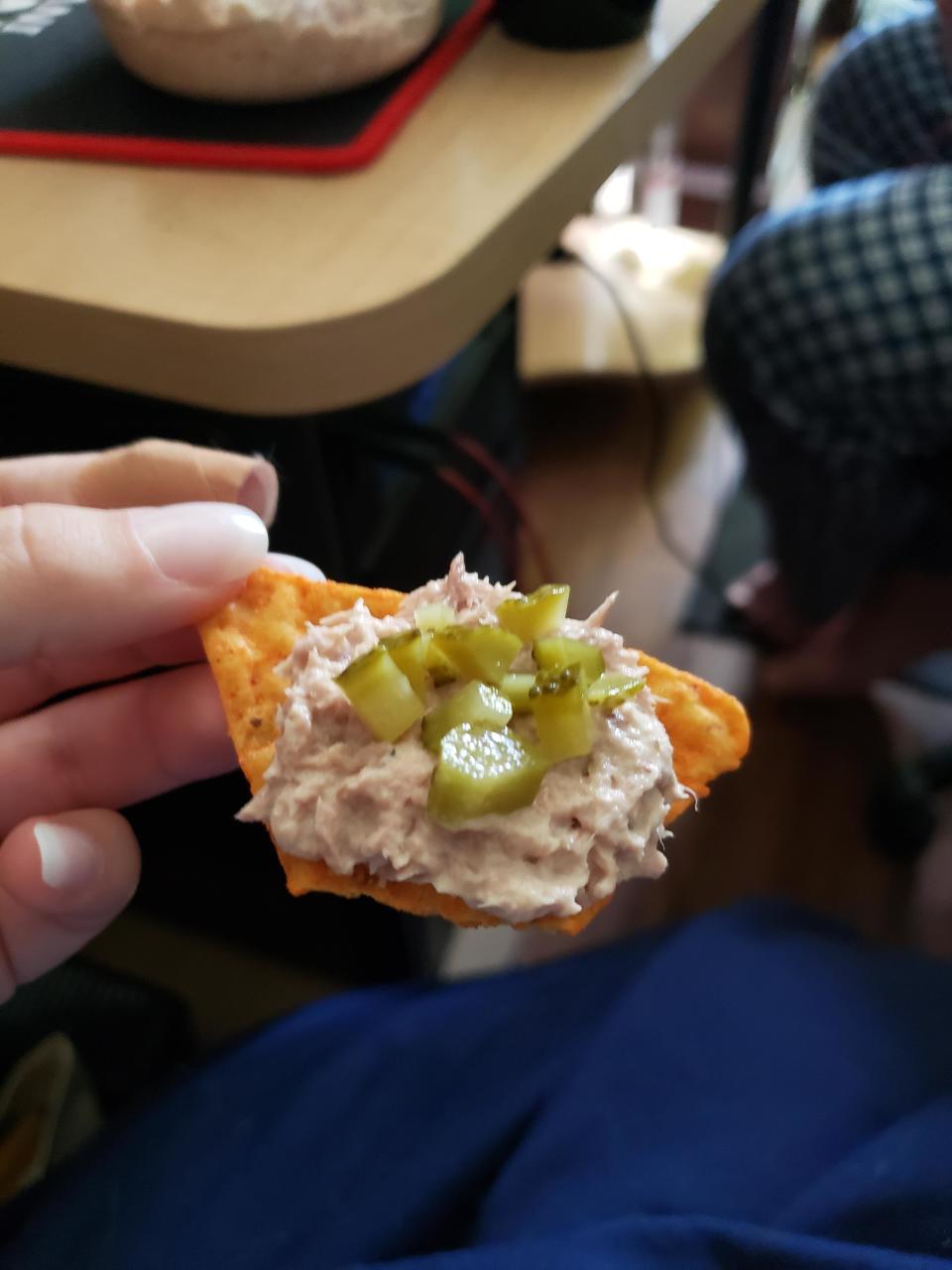 A hand holding a nacho chip topped with a mixture of tuna and pickles, with a table and another person's leg visible in the background