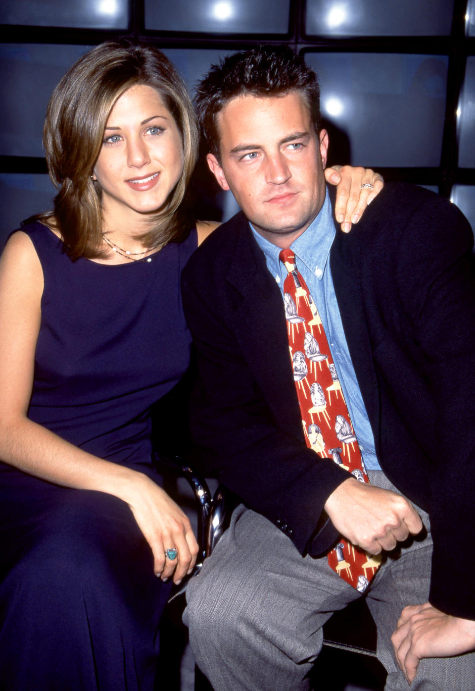 Jennifer Aniston and Matthew Perry pose together. Aniston wears a sleeveless dress. Perry appears in a suit with a character-themed tie