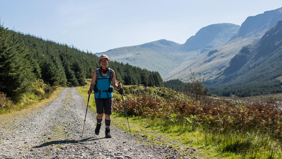 A woman walks along a gravelly hillside, carrying a pair of Black Diamond Pursuit Trekking Poles