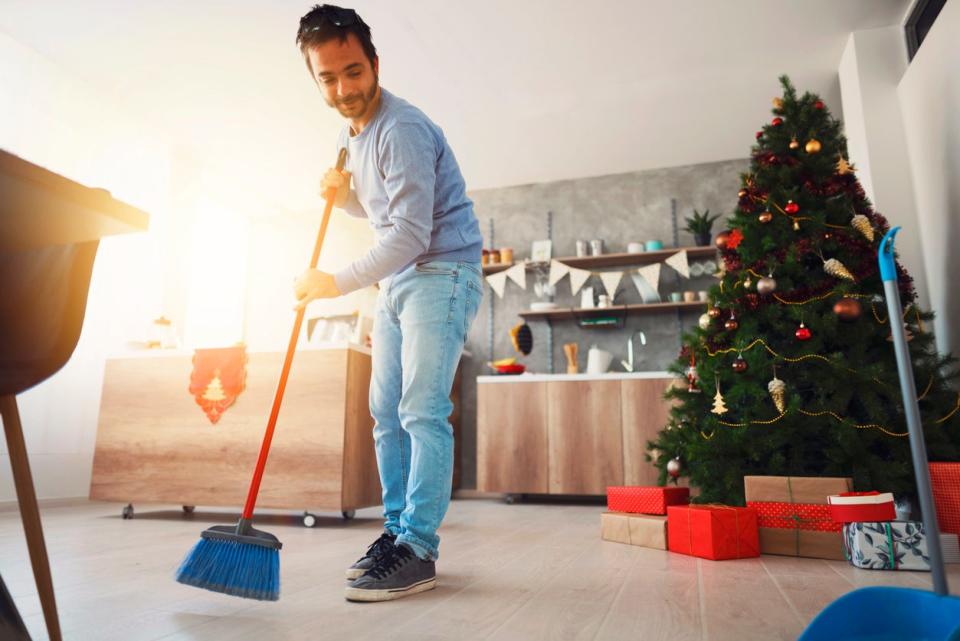 Man sweeping floor with a broom and a dustpan at his home, with a Christmas tree behind him.
