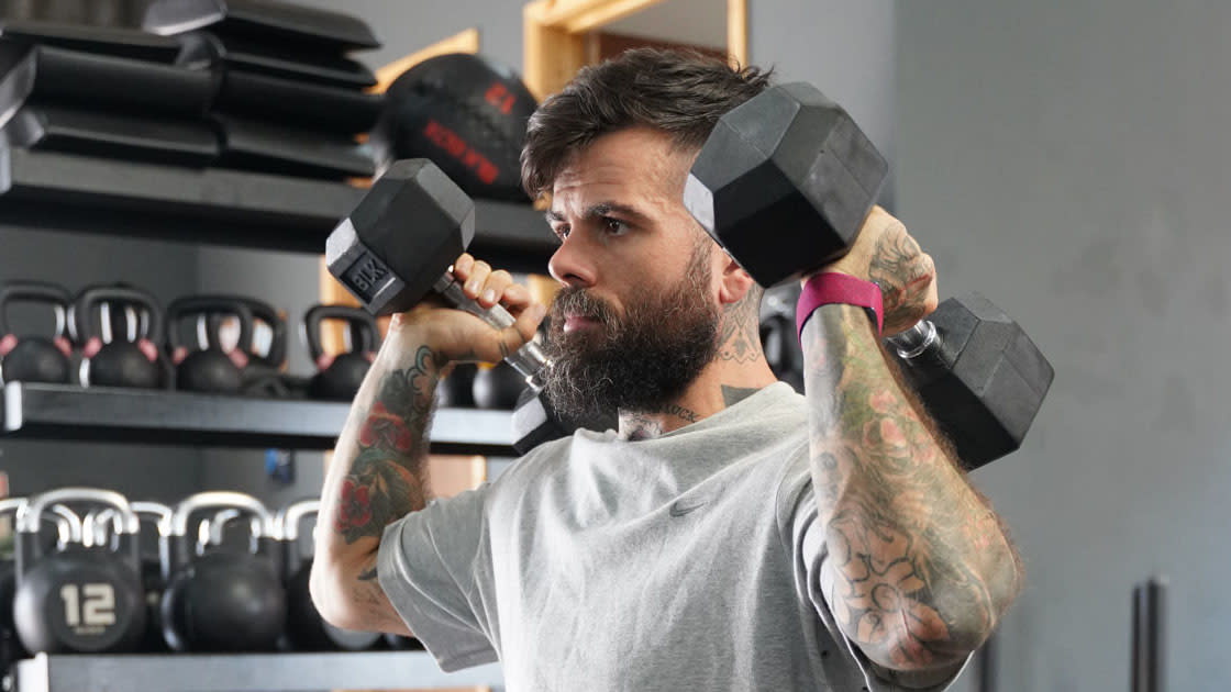  Scott Britton holding dumbbells by his shoulders wearing a pink Whoop tracker on his wrist. 