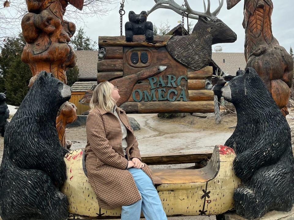 The writer wears a brown coat and blue jeans and sits on a bench with two wood-carved bears on either side and looks up at a sign that says "Parc Omega"