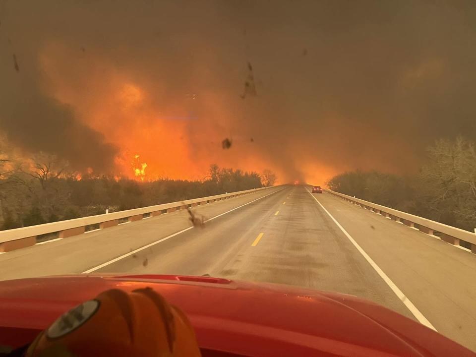 A view of a long stretch of a nearly empty road with the Smokehouse Creek Fire raging in the distance.