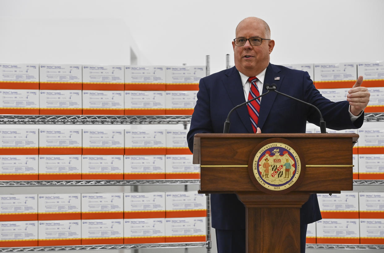 Gov. Larry Hogan makes a point in front of two racks packed with rapid screening tests for COVID.