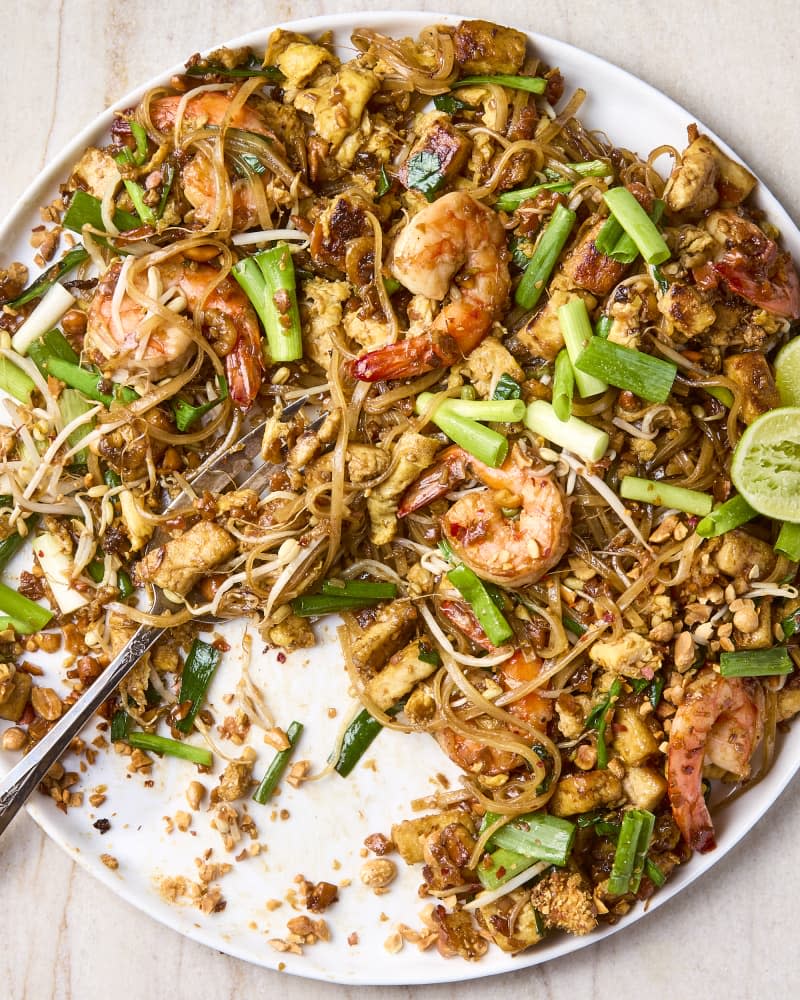 overhead shot of pad thai on a large white round plate, garnished with lime halves and green onion, with a serving taken from the plate