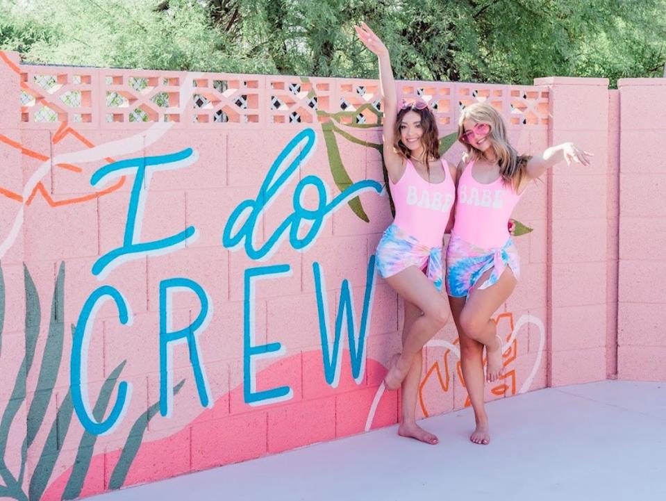 Zwei Frauen in rosa Badeanzügen stehen vor einem Wandgemälde mit der Aufschrift "I do crew". Quelle:The Femme House - Copyright: The Femme House