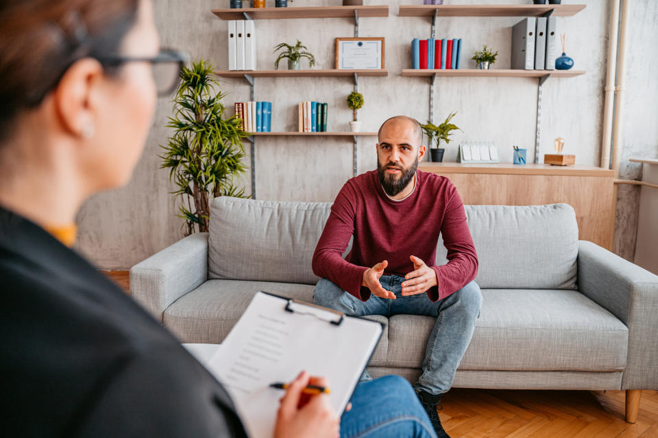 Man in a casual sweater and jeans sitting on a sofa, gesturing while talking to a person holding a notepad