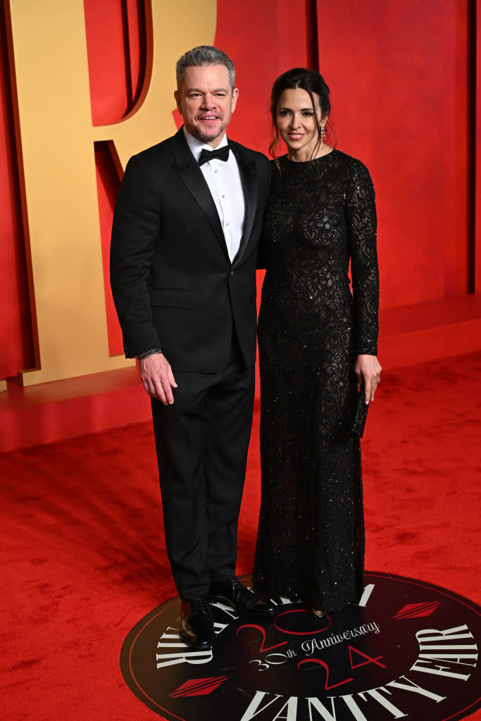 Two individuals posing at Vanity Fair event; one in a tuxedo, the other in a long-sleeved, embellished gown