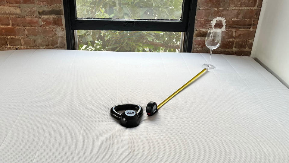 Photo of Helix Midnight mattress in a bedroom, with a weight on it, and a wine glass stood upright on the mattress surface nearby. A tape measure shows the distance between glass and weight.