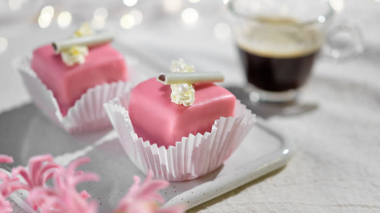 Two pink petit fours on a plate with coffee in background