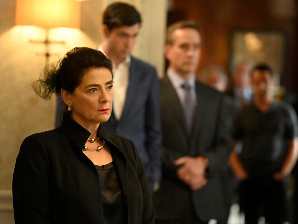 An older woman wearing all black stands in an ornate living room with a stern expression on her face.