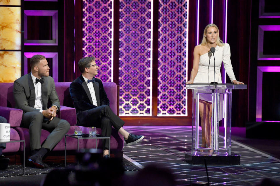 Nikki stands at a podium with two other celebrities sitting on stage