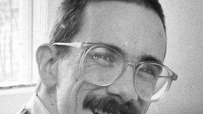 bill watterson smiles at the camera while looking over one shoulder, he wears large circular glasses a collared shirt and a sweater