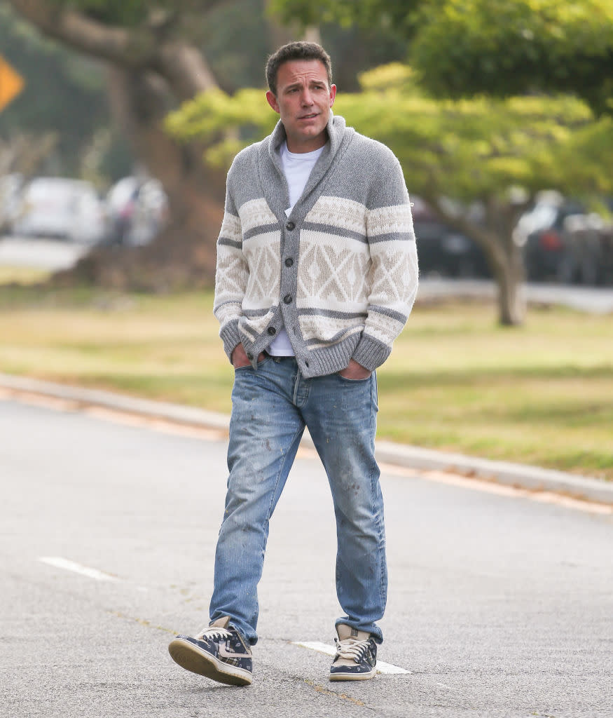 Man walking outside on a road, wearing a patterned cardigan, white shirt, blue jeans, and sneakers