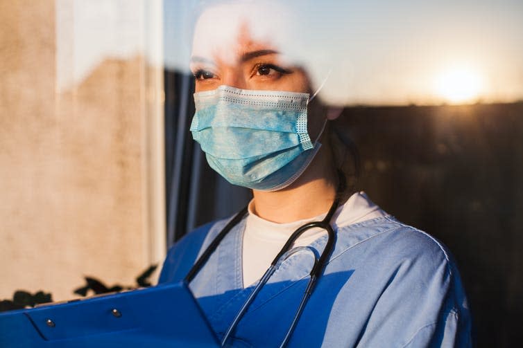 A carer wearing a mask and holding a clipboard looks out of a window.