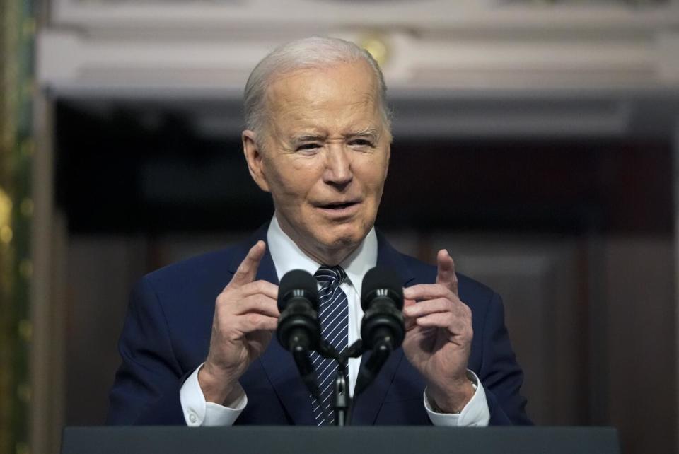 A man in a dark suit and dark striped tie gestures with his hands while speaking before microphones